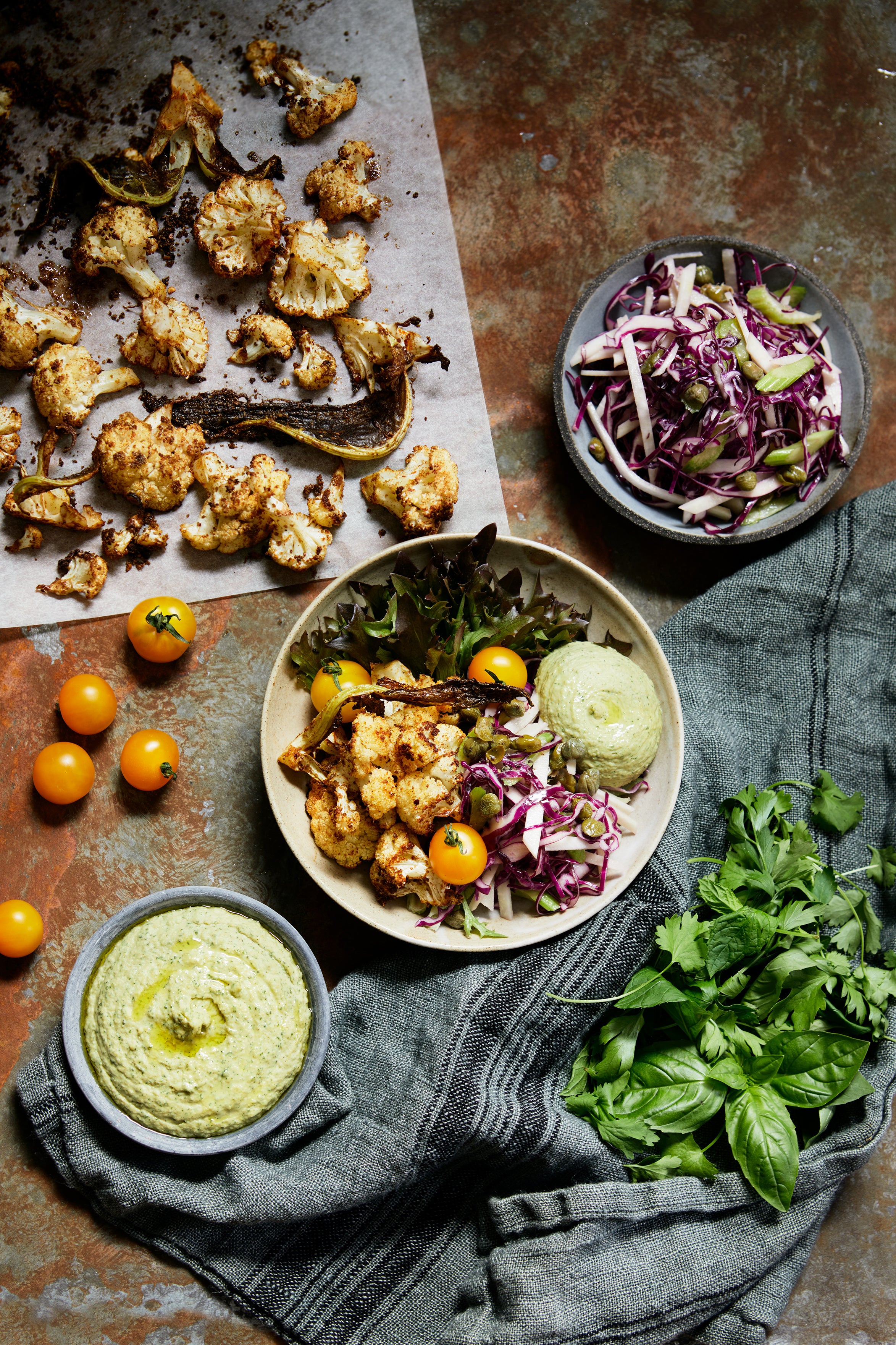 skålar med rostad blomkål, rödkål, tomat med mera fotade ovan ifrån på ett rostigt underlag. Runtom en kökshandduk med färska örter på och ett bakplåtspapper med rostad blomkål på. 